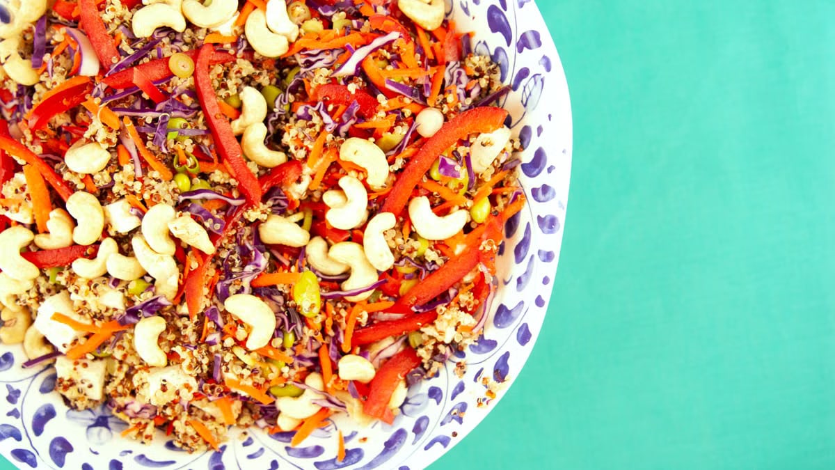 Colorful quinoa salad with cashews, peppers, and cabbage in a decorative bowl on a turquoise background.